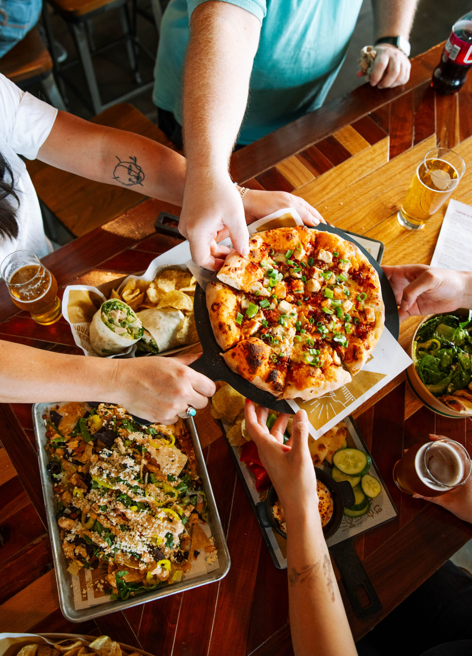 Four people reach for a shared pizza at a Boulevard Brewing Co. restaurant table also covered with a tray of nachos, a wrap, a side salad, and drinks.