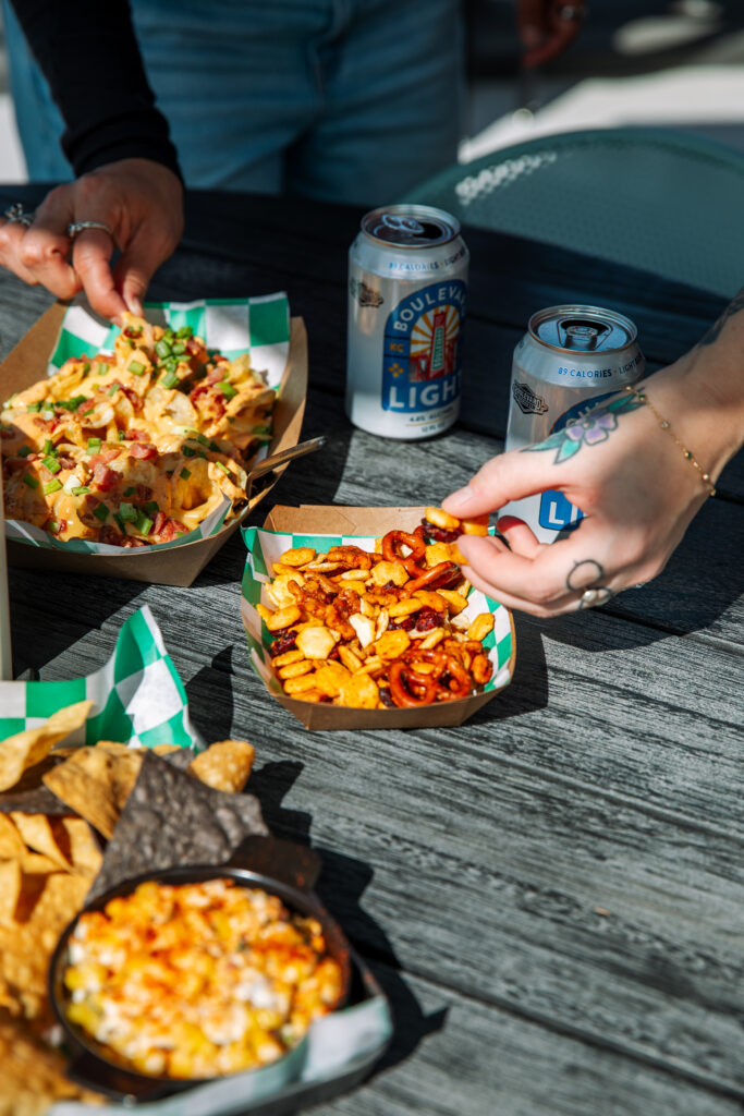 Two hands reach for snacks, including a tray of loaded nachos, a bowl of snack mix, and a skillet of queso, set on an outdoor table at the Boulevard Brewing Co. Beer Hall next to two cans of Boulevard Light beer.