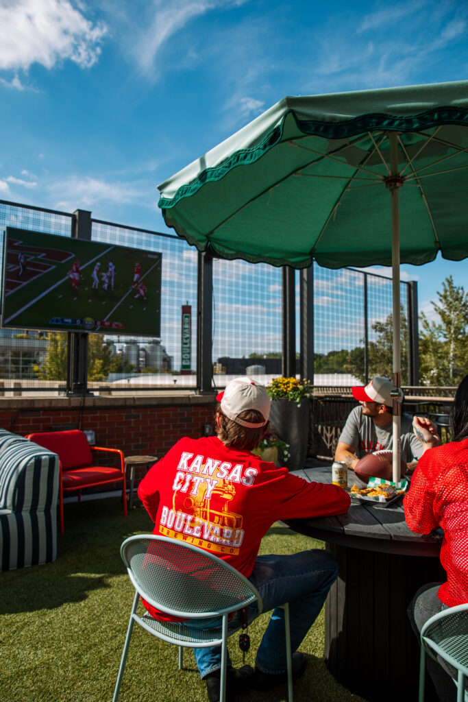 Three people sit at a round table under a green patio umbrella at the Boulevard Brewing Co. Beer Hall, watching a football game on a large outdoor screen.