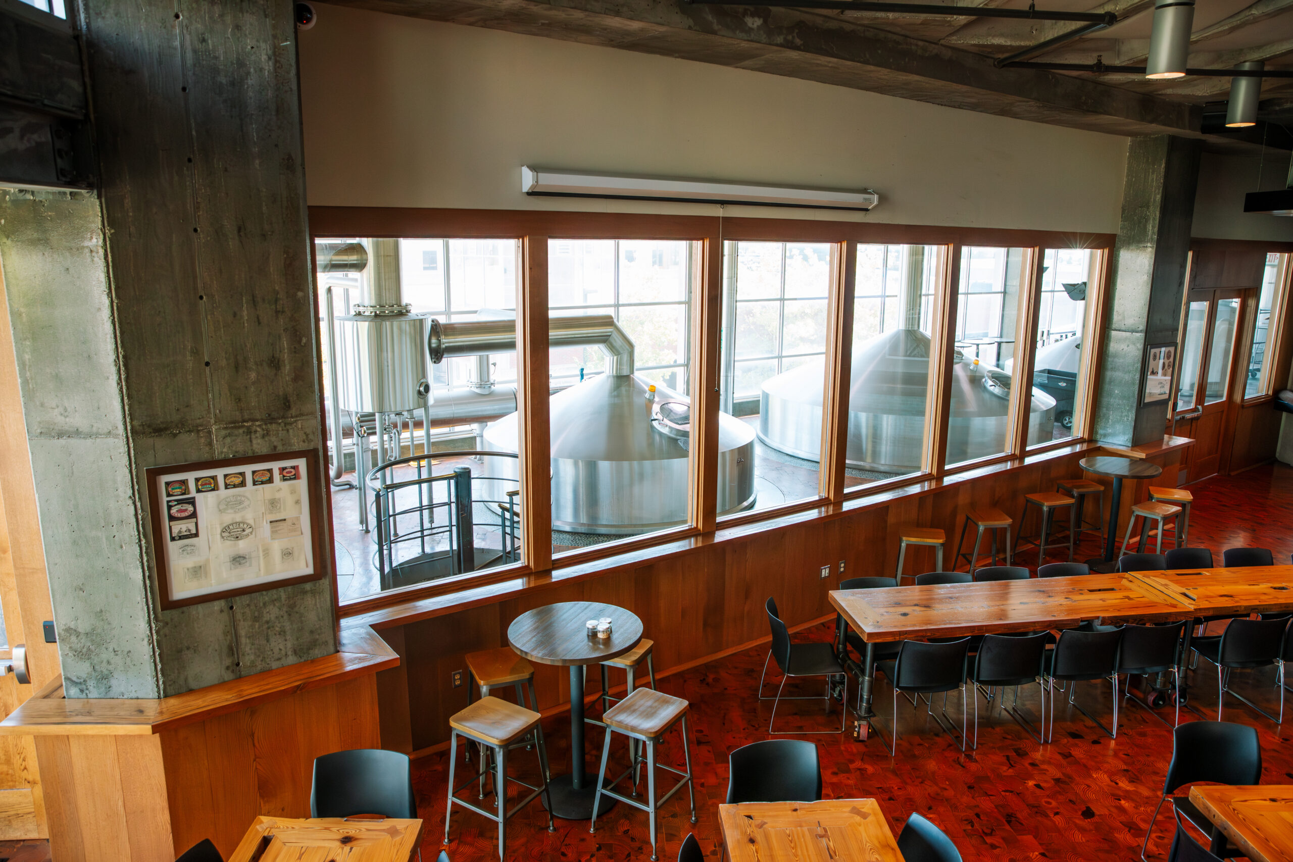 A brewery taproom with wooden tables and black chairs, large interior windows offering a view of stainless steel brewing tanks and equipment in the production area. Natural light fills the modern, industrial-style space.