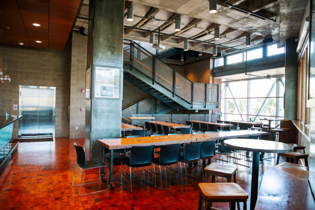 A modern, spacious cafeteria with wooden tables, black chairs, large windows letting in natural light, a metal staircase, and exposed concrete walls and ceiling. The space appears clean and empty.