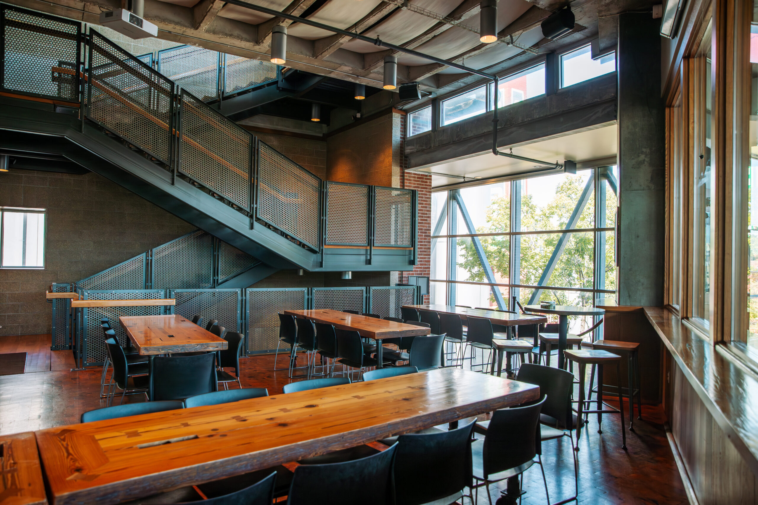 Modern industrial-style dining area with wooden tables, black chairs, large windows letting in natural light, and a metal staircase leading to an upper level. The space features exposed ceilings and brick walls.