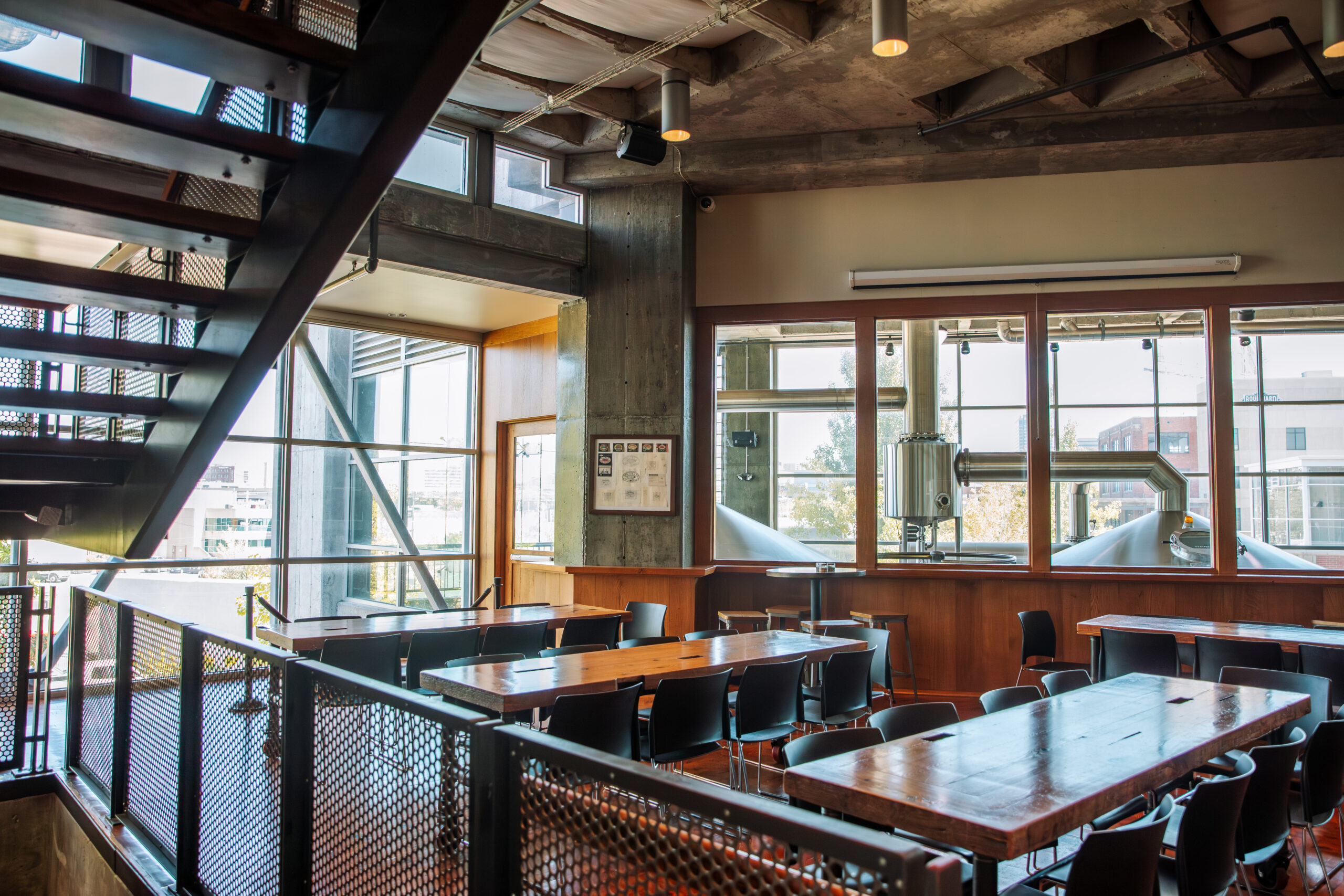 A modern industrial-style room with long wooden tables, black chairs, large windows, metal stairs, and exposed concrete and pipes, filled with natural light.
