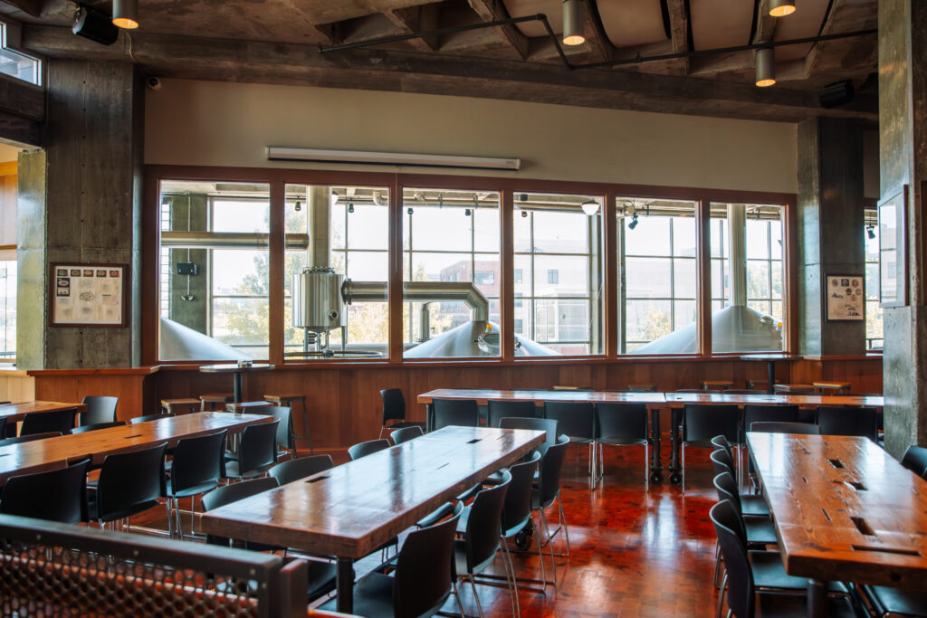 A spacious, modern industrial dining area with wooden tables and black chairs, large windows, and visible metal brewing equipment outside. Natural light fills the room, highlighting the polished wood floor.