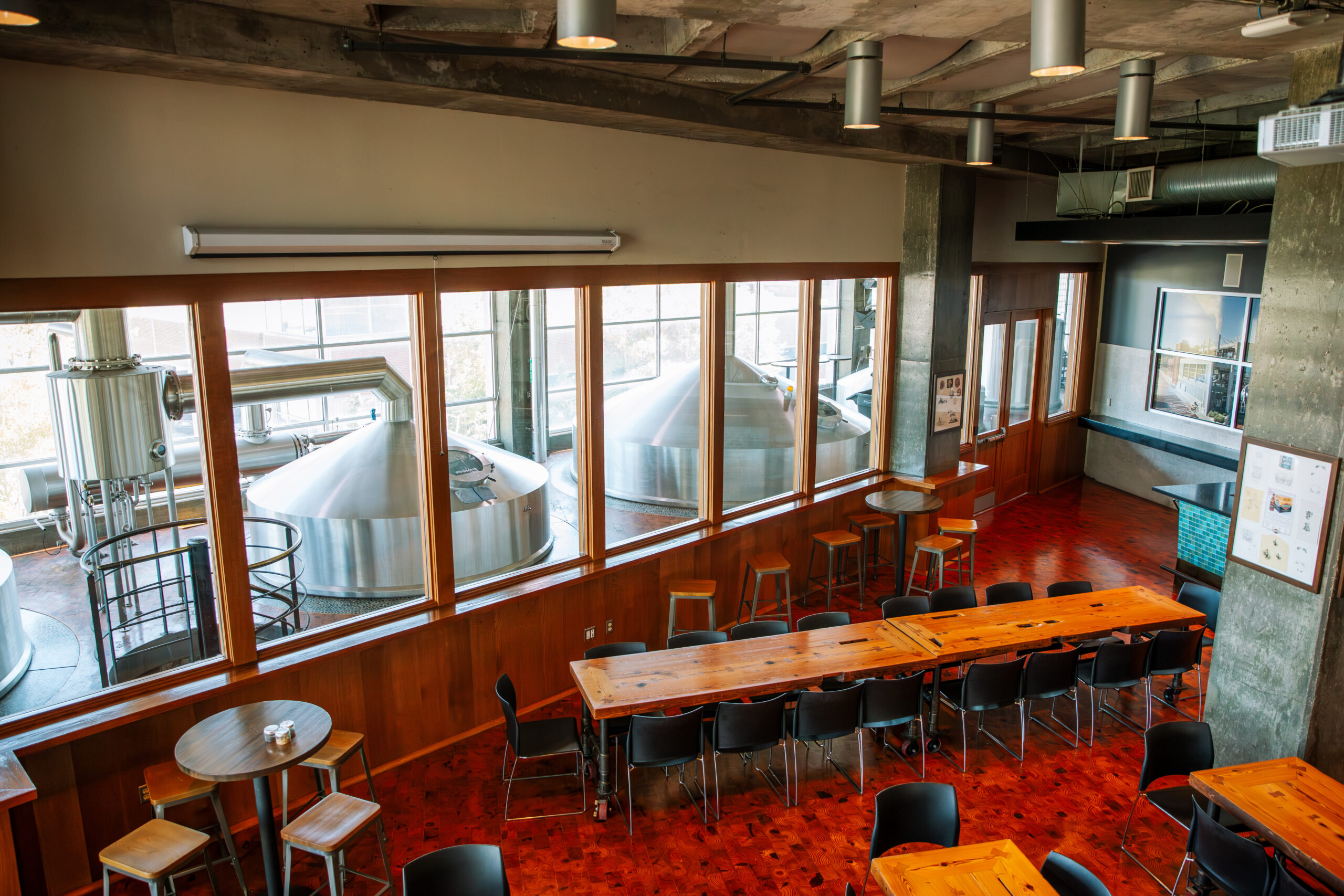 A modern brewery interior with large steel brewing tanks visible through glass windows, long wooden tables and chairs arranged for guests, and a polished red floor. The space is empty and well-lit.