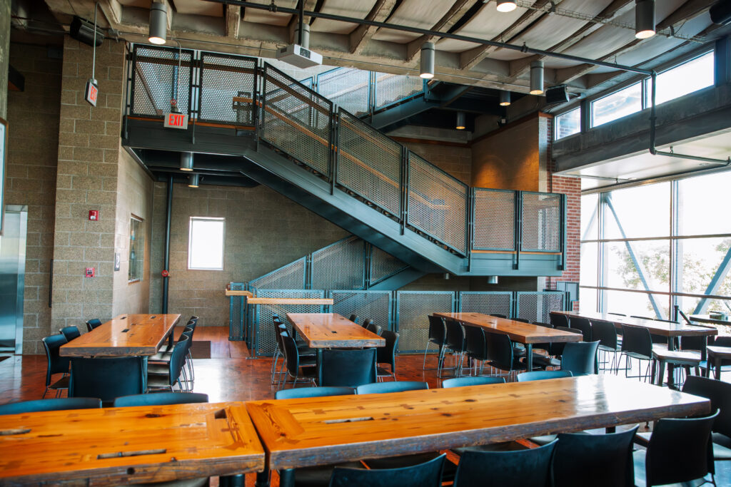 Modern cafeteria with wooden tables, black chairs, and large windows letting in natural light. An industrial-style metal staircase is in the background, and the space has exposed ceilings and brick walls.