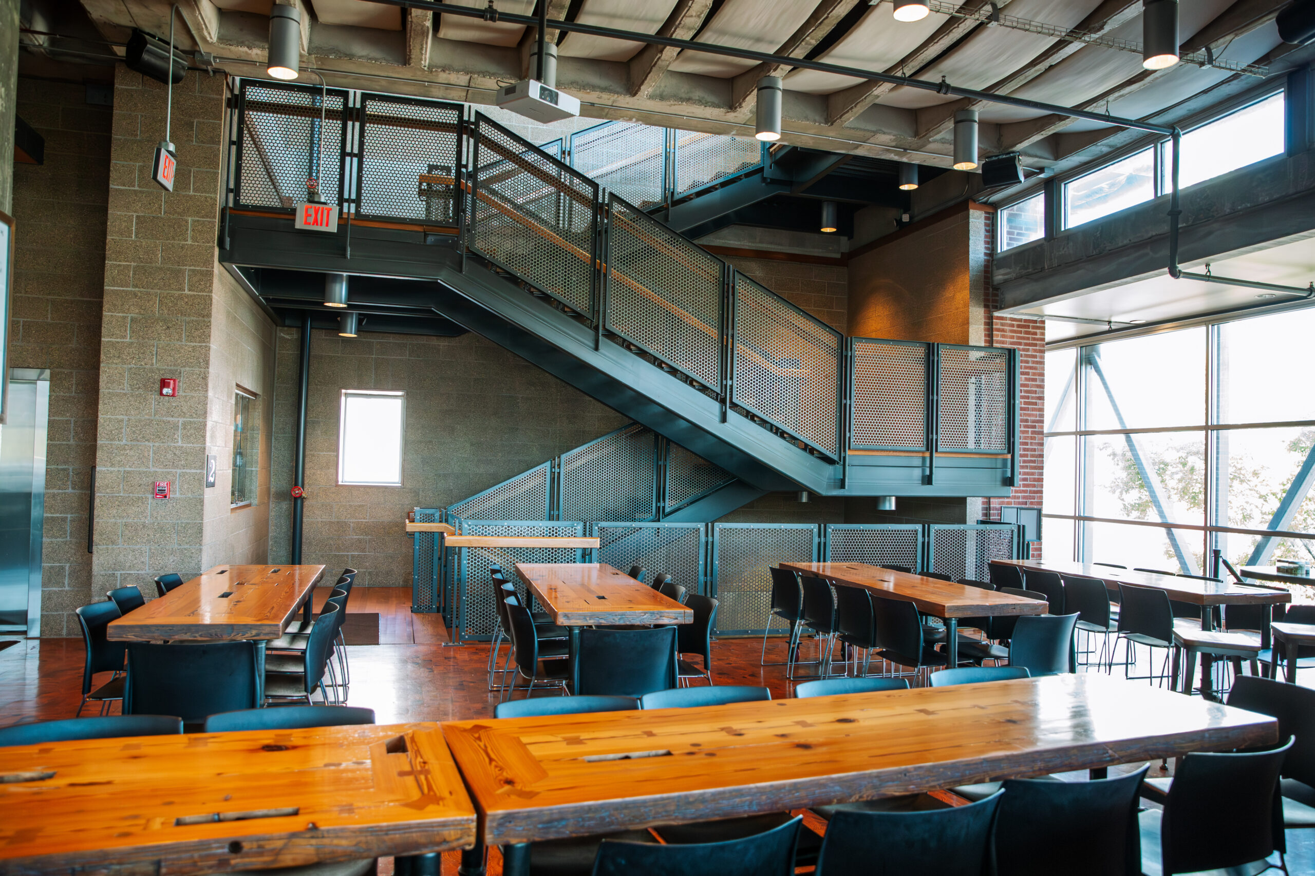 Modern cafeteria with wooden tables, black chairs, and large windows letting in natural light. An industrial-style metal staircase is in the background, and the space has exposed ceilings and brick walls.