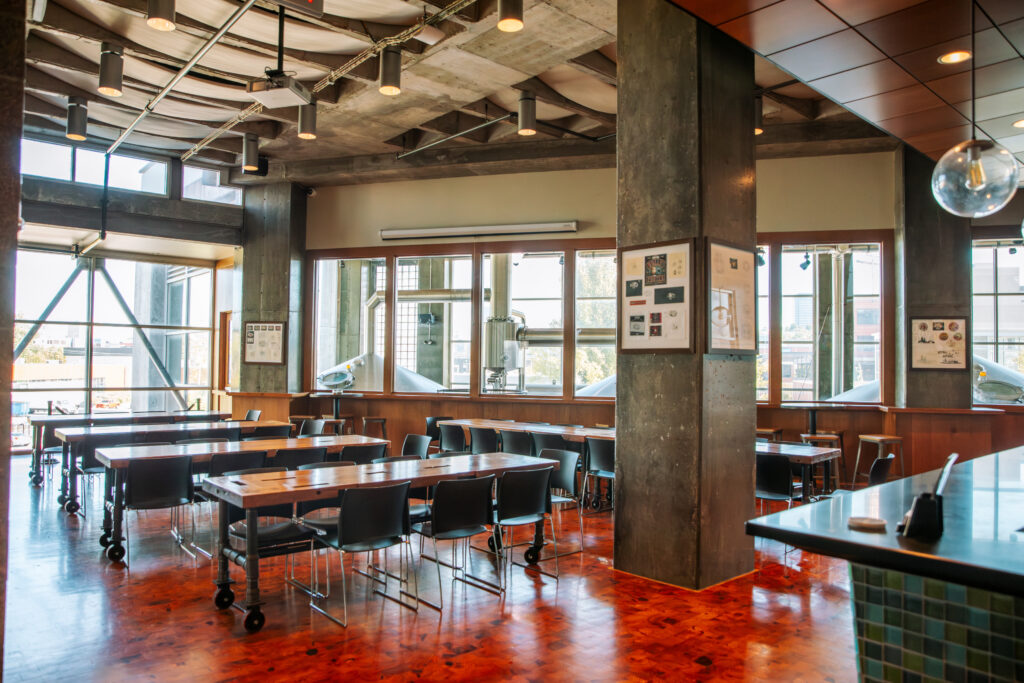 A modern, spacious cafe or dining area with wooden floors, large windows letting in natural light, industrial-style ceilings, and tables with black chairs arranged neatly.
