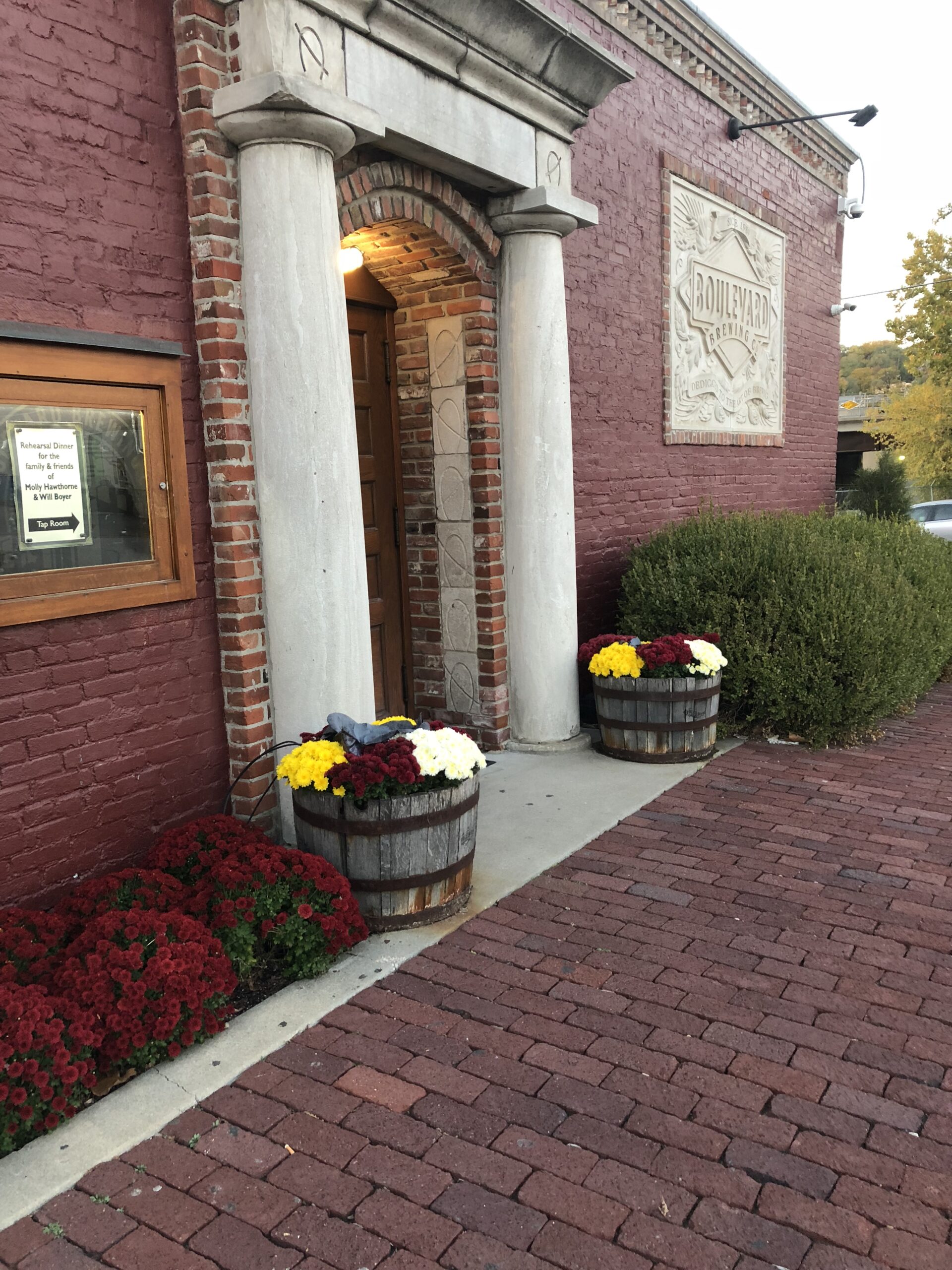 A brick building with a stone column entrance, flanked by barrels filled with colorful flowers. Red flowers and green bushes line the path, and a sign reading "Boulevard" is mounted on the wall.
