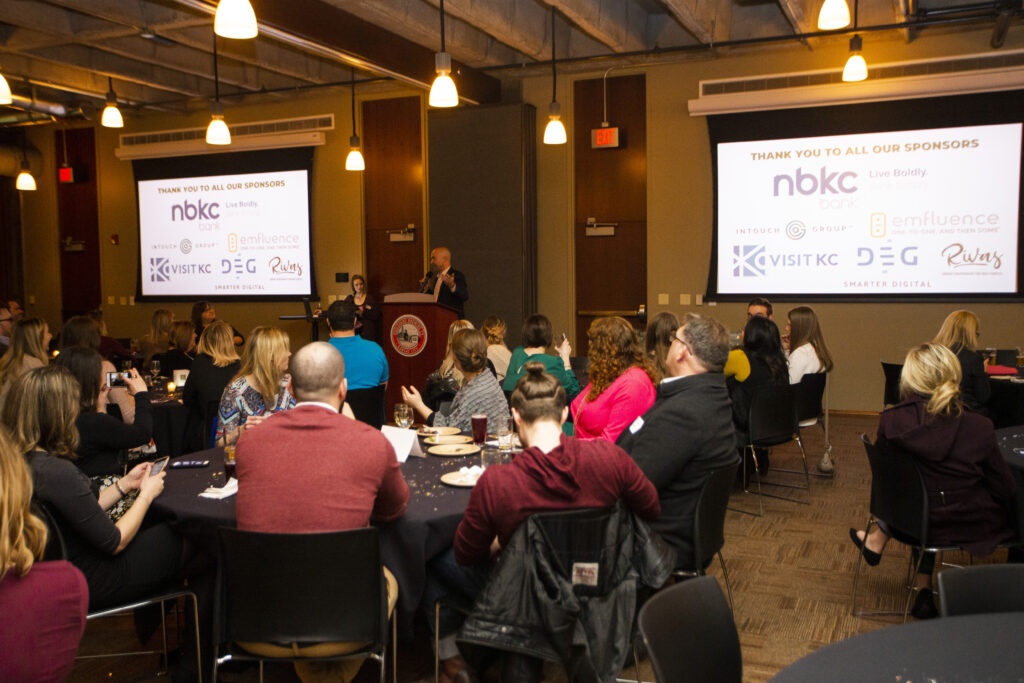 People seated at round tables in a conference room listen to a speaker at a podium, with a large screen behind displaying sponsor logos and a thank you message.