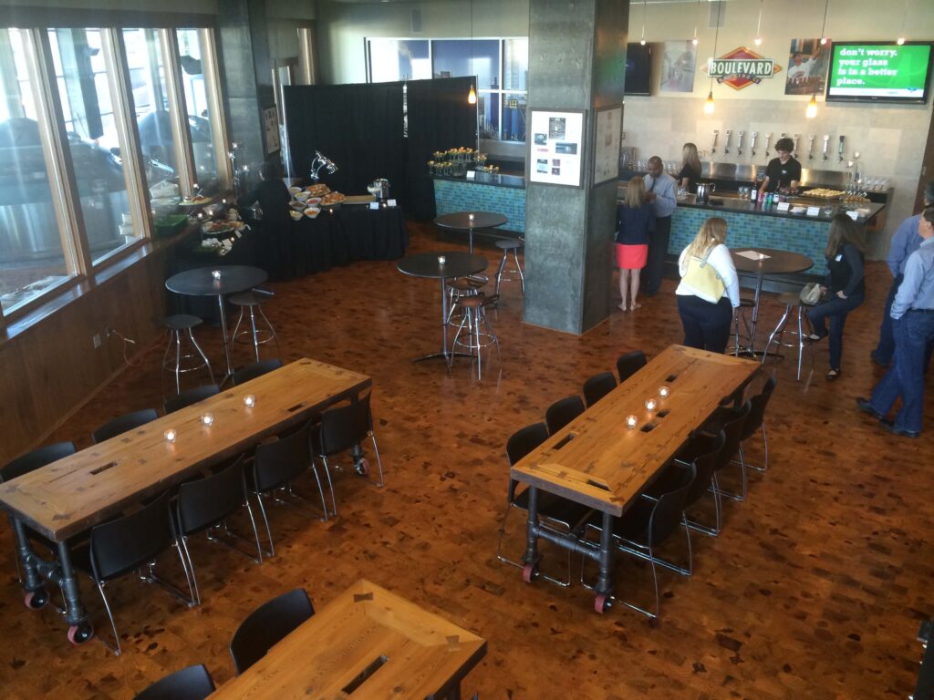Spacious modern room with wooden tables and chairs, a few people near a bar area, and a buffet set up in the corner. The floor is wood, and candles are on the tables. Large windows line one wall.