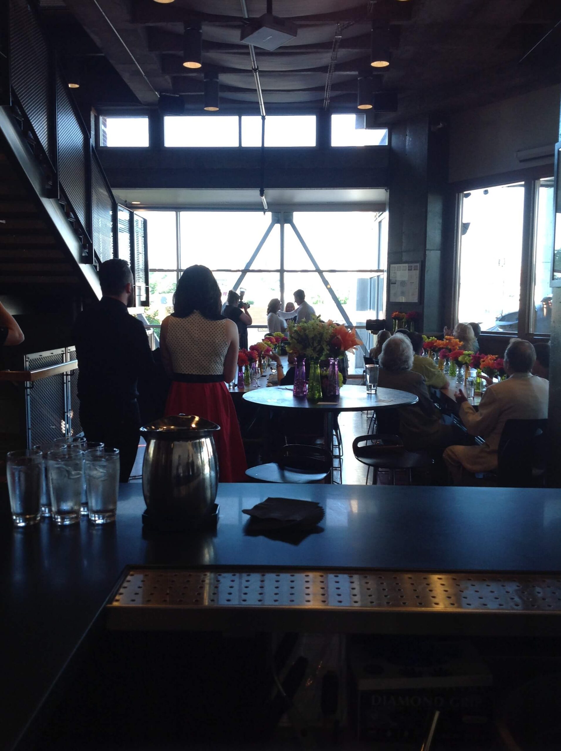 A group of people gather in a modern, industrial-style room with large windows. Several round tables hold colorful flower arrangements, and some guests are seated while others stand and socialize.