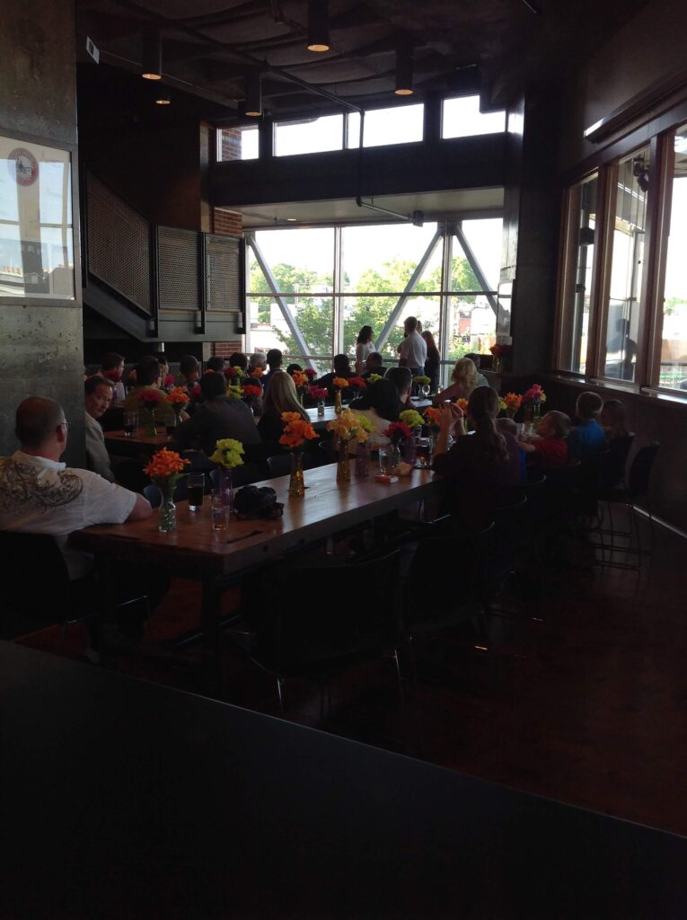 People are seated around a long table in a modern, sunlit restaurant. The table is decorated with colorful flower arrangements, and large windows let in natural light from outside.