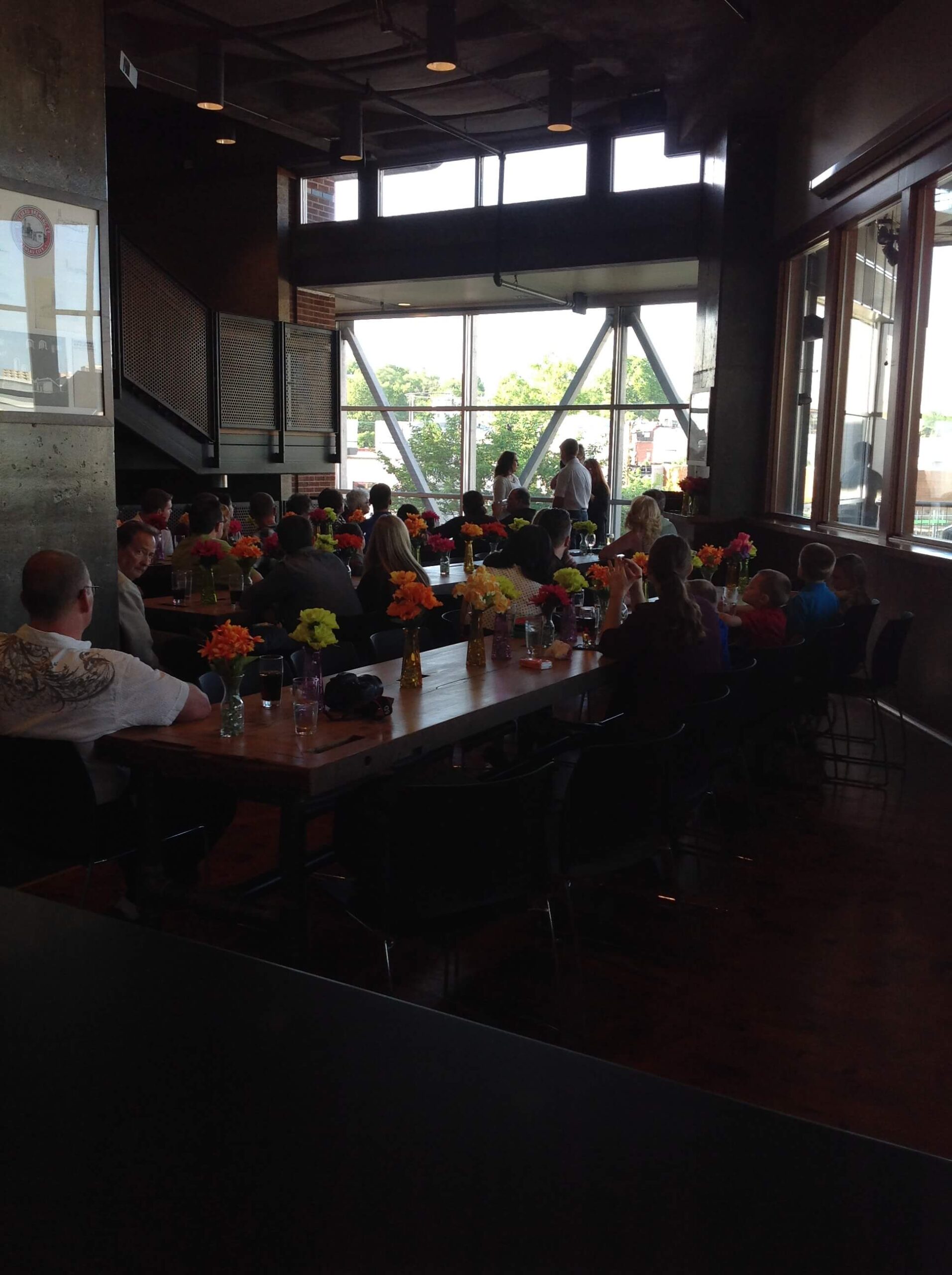 People are seated around a long table in a modern, sunlit restaurant. The table is decorated with colorful flower arrangements, and large windows let in natural light from outside.