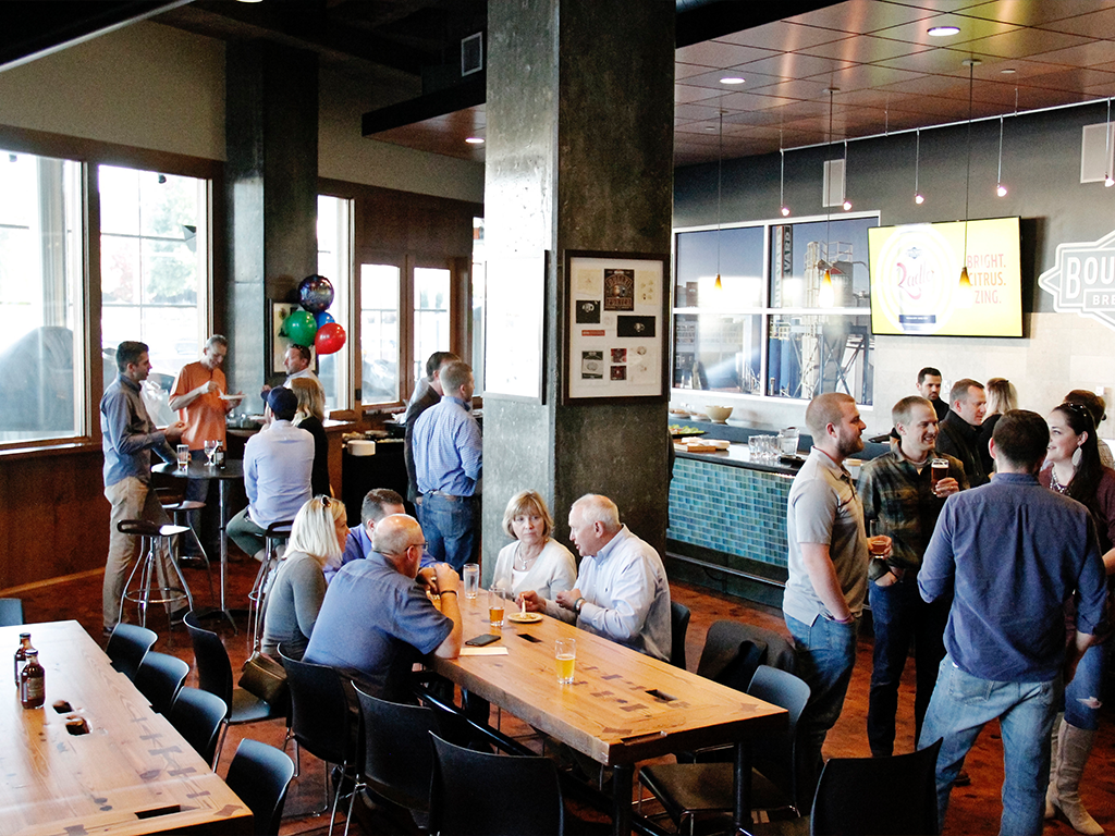 A group of people socializing in a modern bar. Some are standing with drinks, while others sit at wooden tables. Bright windows light the space, and colorful balloons are in the background. A wall-mounted TV displays a logo on the right side.