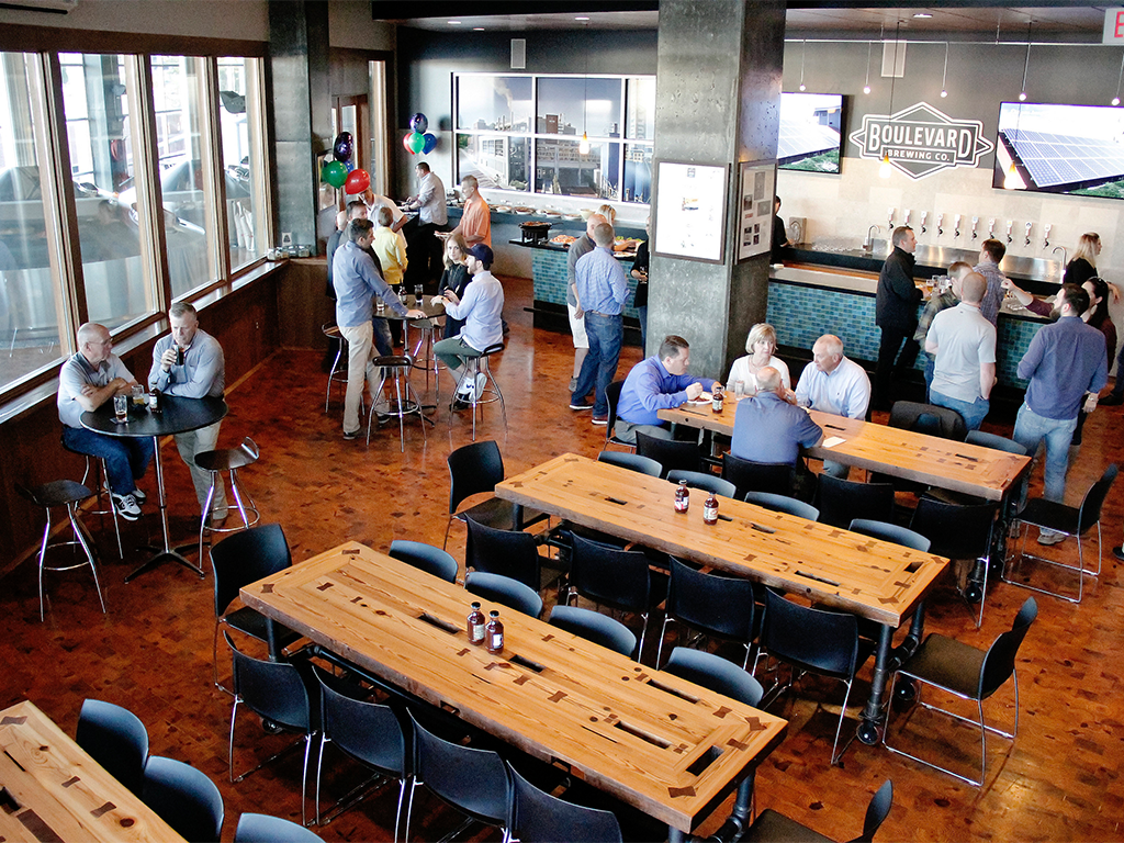A lively brewery taproom with people gathered at wooden tables, talking and enjoying drinks. Some are standing by the bar, and a few are seated in groups. The room features large windows, festive balloons, and a TV displaying the Boulevard Brewing logo.
