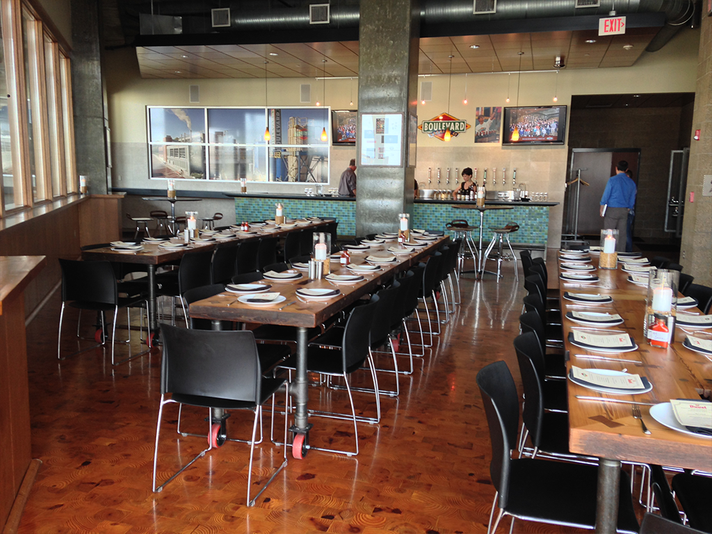 A modern restaurant interior with long wooden tables set with plates and utensils. The counter area features a display of beer taps and wall art. Two people are visible near the counter, and the space has industrial-style decor elements.