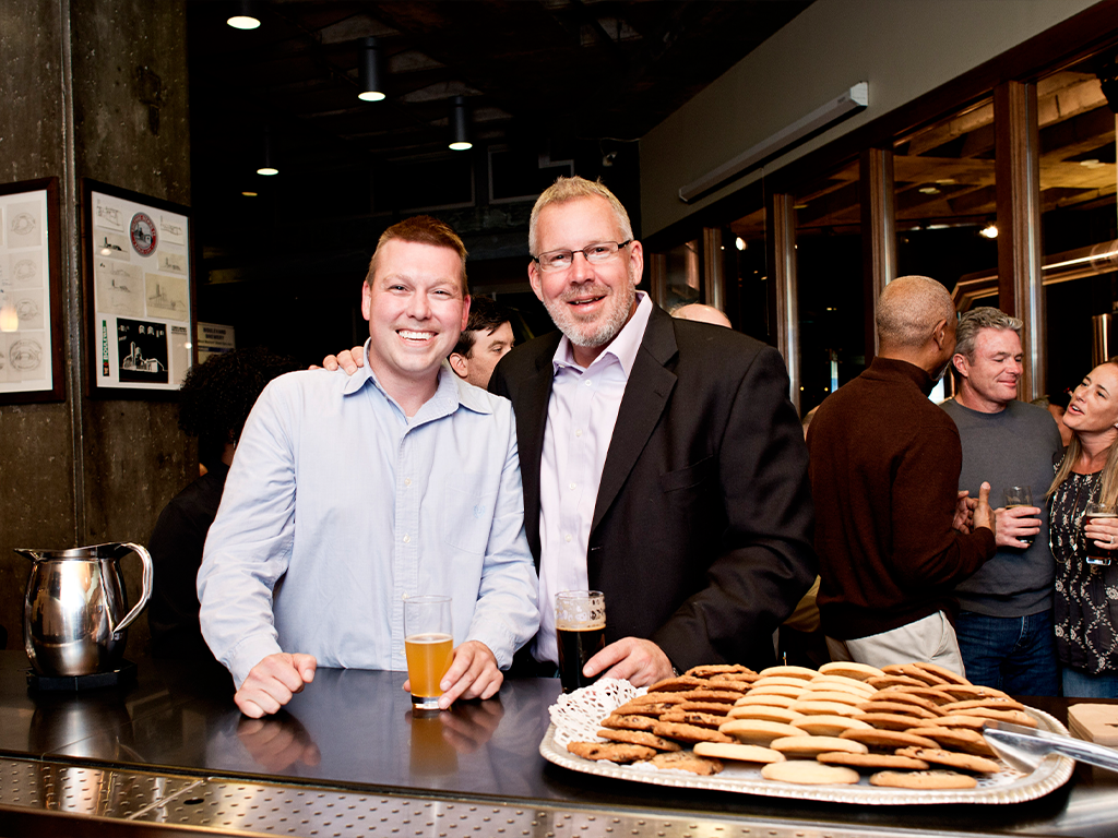 Two men are smiling and standing side by side at a bar. One holds a glass of beer, and the other also holds a drink. A tray of cookies is on the counter in front of them. Several people are socializing in the background.