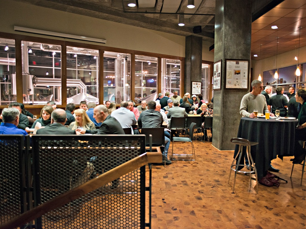 People are gathered in a bustling, warmly lit brewery or restaurant. Groups sit at tables, engaged in conversation. A man stands at a high-top table with drinks. Large windows reveal brewing equipment in the background.