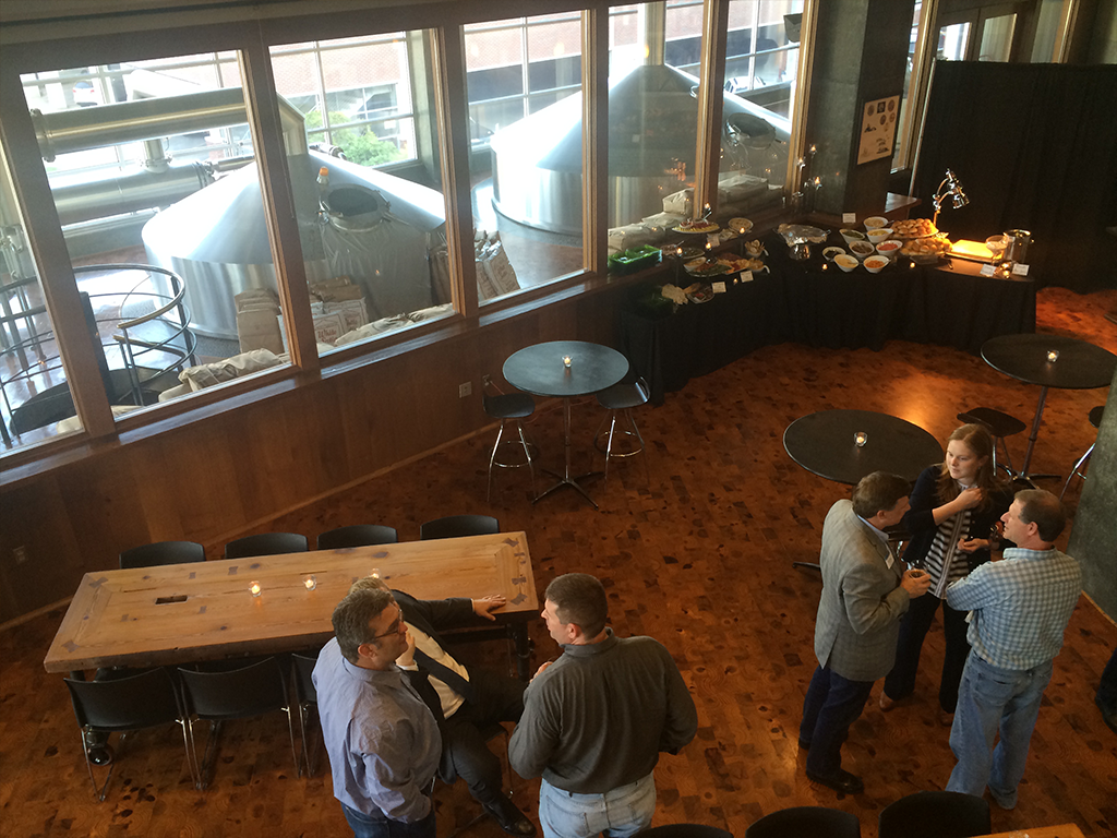 A brewery event space featuring fermentation tanks visible through large windows. Three men sit at a wooden table, while a man and a woman converse nearby. Round, black tables and a buffet with various dishes are set up for guests.