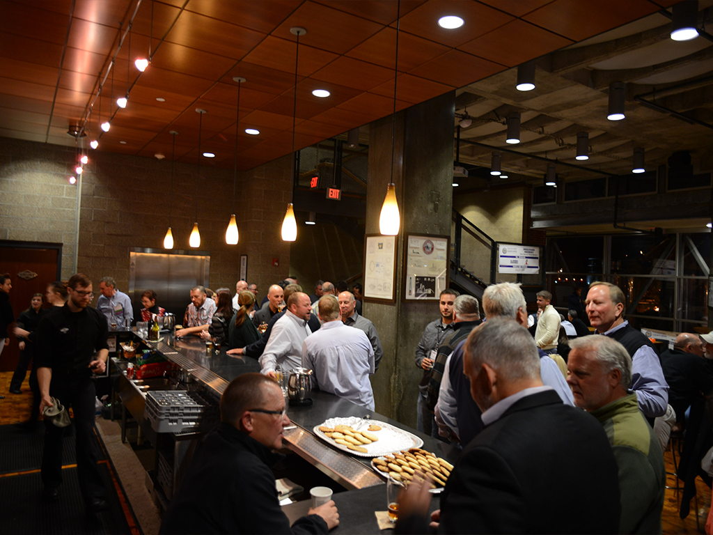 People are gathered in a modern bar with low lighting, enjoying drinks and conversation. A bartender serves behind the counter, while a tray of appetizers is visible. The setting is lively and social with a warm atmosphere.