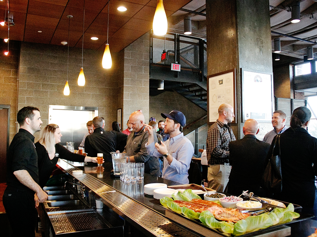 A group of people socializing at a modern, lively bar. Several individuals are engaged in conversation near the counter, which displays a platter of food. The ambiance is warm with hanging lights and contemporary decor.