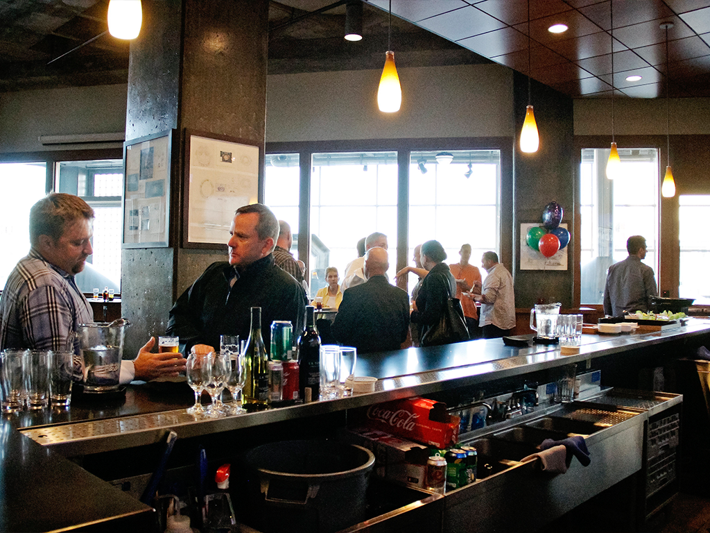A modern bar setting with several people standing and conversing. The counter is stocked with glasses and various drinks. Large windows in the background let in natural light, and balloons add a festive touch to the room.