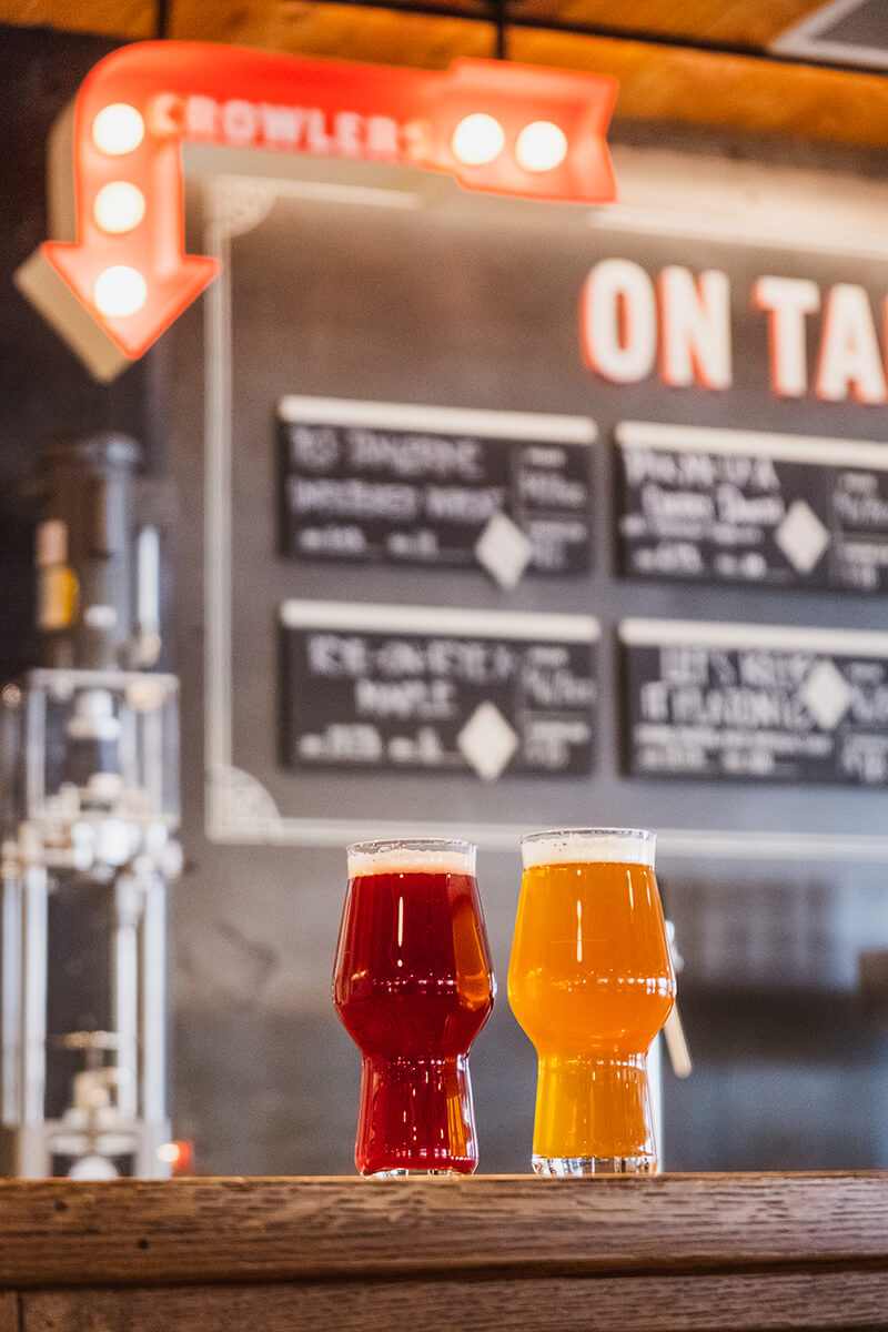 Two pint glasses containing red and gold beer sit on a wooden counter in a bar. Behind them, a menu board displays various beers on tap, with a bright, arrow-shaped sign pointing downwards. The background features industrial-style decor.