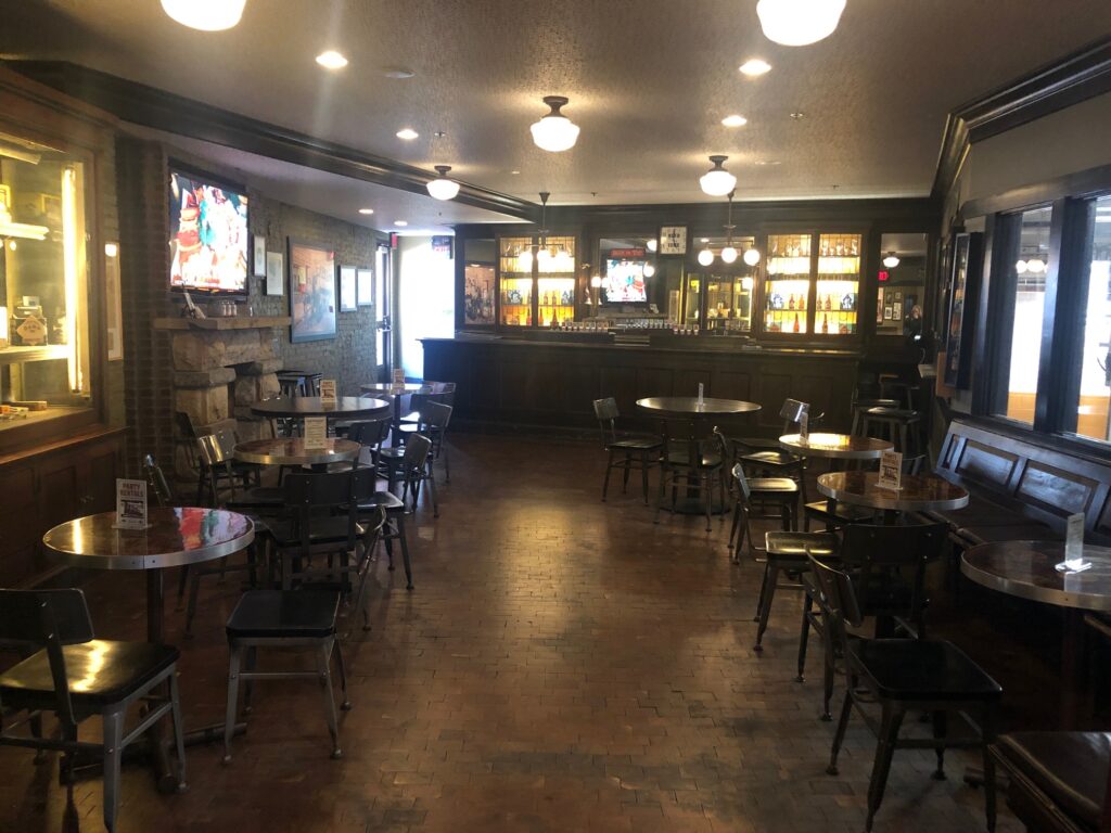 A warmly lit, empty bar with wooden tables and chairs, brick flooring, a long counter at the back, and TVs on the walls. Shelves behind the bar display bottles, and large windows line the right side.