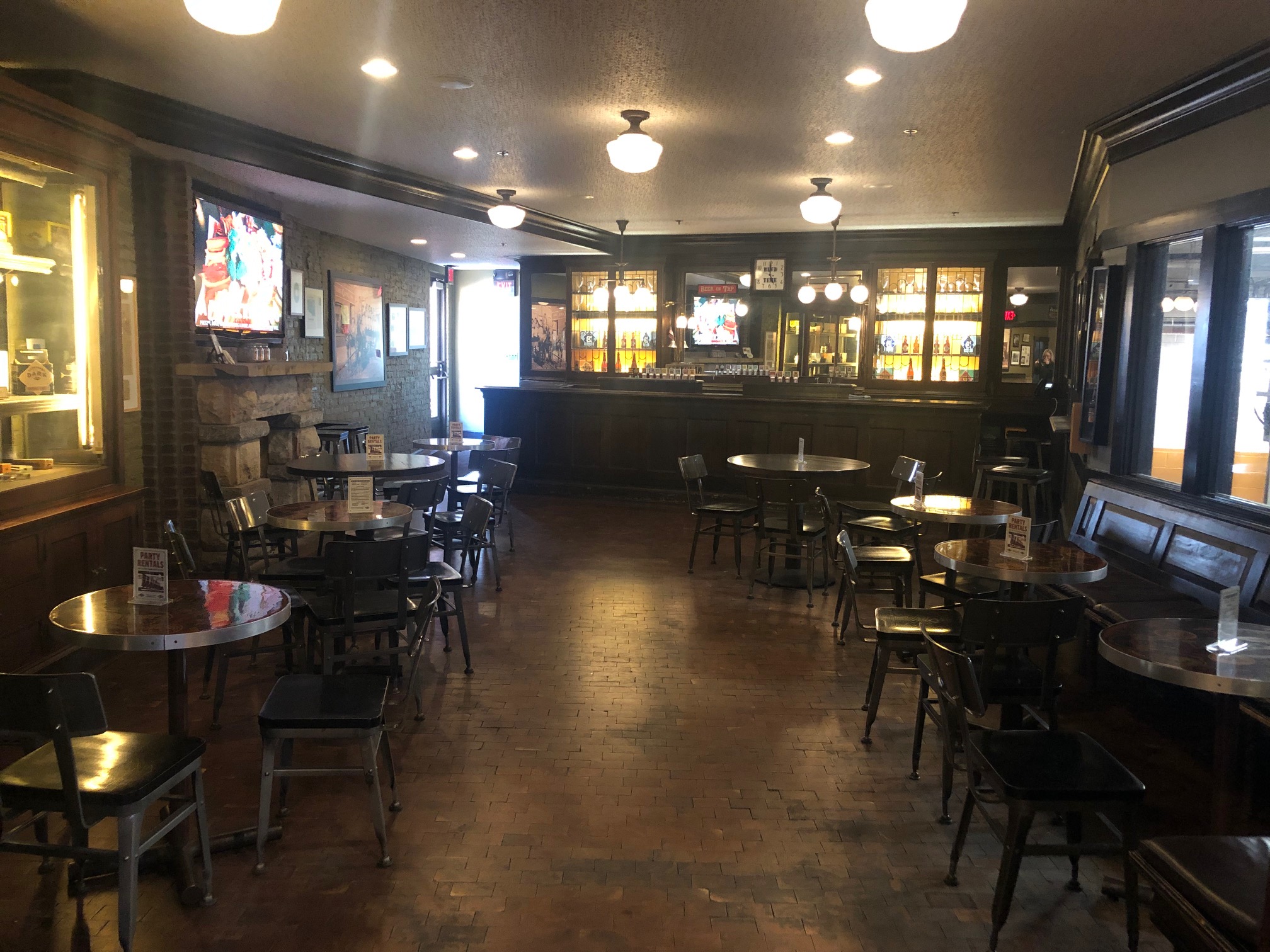 A warmly lit, empty bar with wooden tables and chairs, brick flooring, a long counter at the back, and TVs on the walls. Shelves behind the bar display bottles, and large windows line the right side.