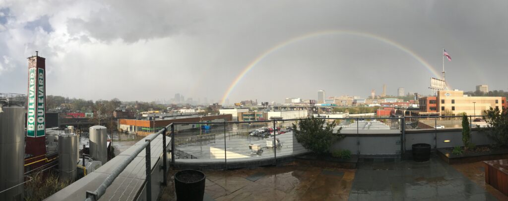 A panoramic view of an urban rooftop with wet pavement, overlooking city buildings and a brewery tank. A faint rainbow arches across a cloudy sky, and an American flag waves on a pole to the right.