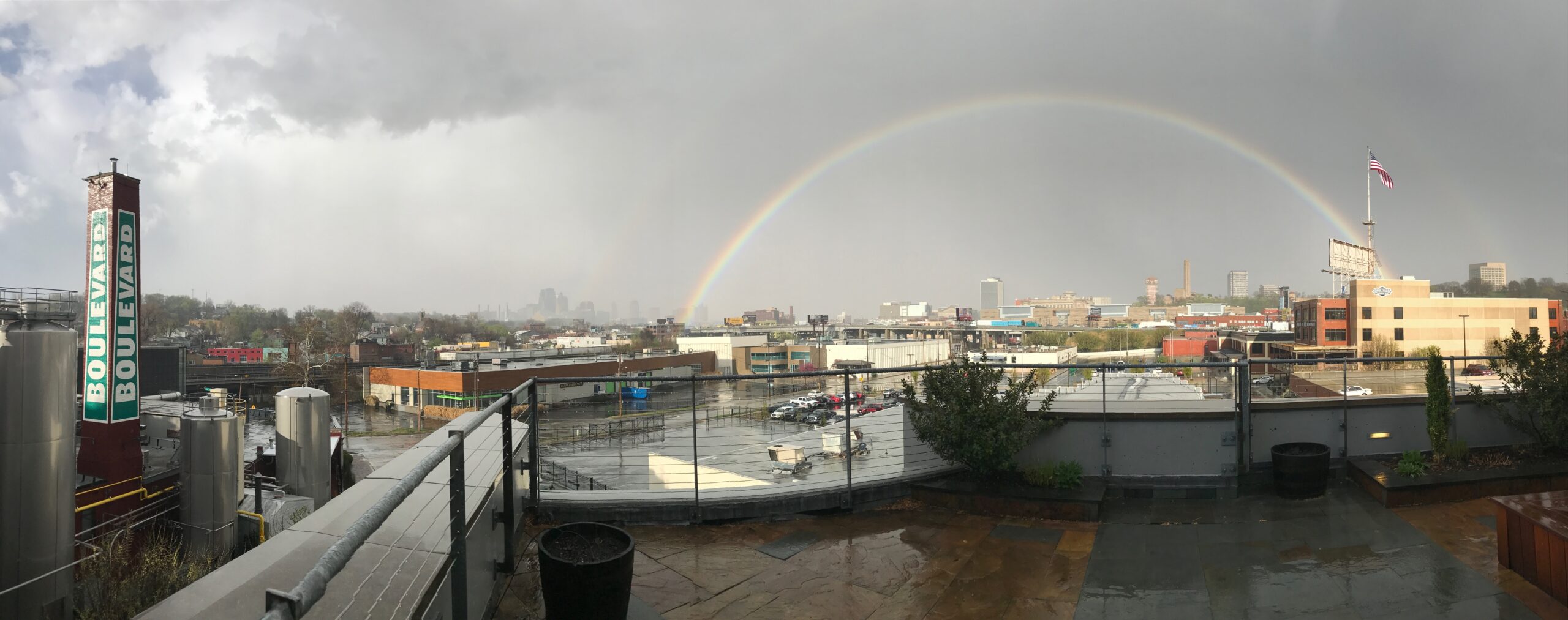 A panoramic view of an urban rooftop with wet pavement, overlooking city buildings and a brewery tank. A faint rainbow arches across a cloudy sky, and an American flag waves on a pole to the right.