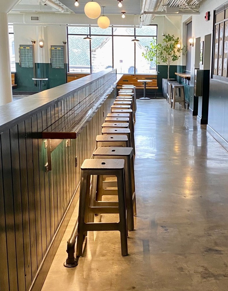 A row of wooden bar stools lines a long counter in a modern, well-lit café with polished concrete floors, large windows, and minimalistic decor.