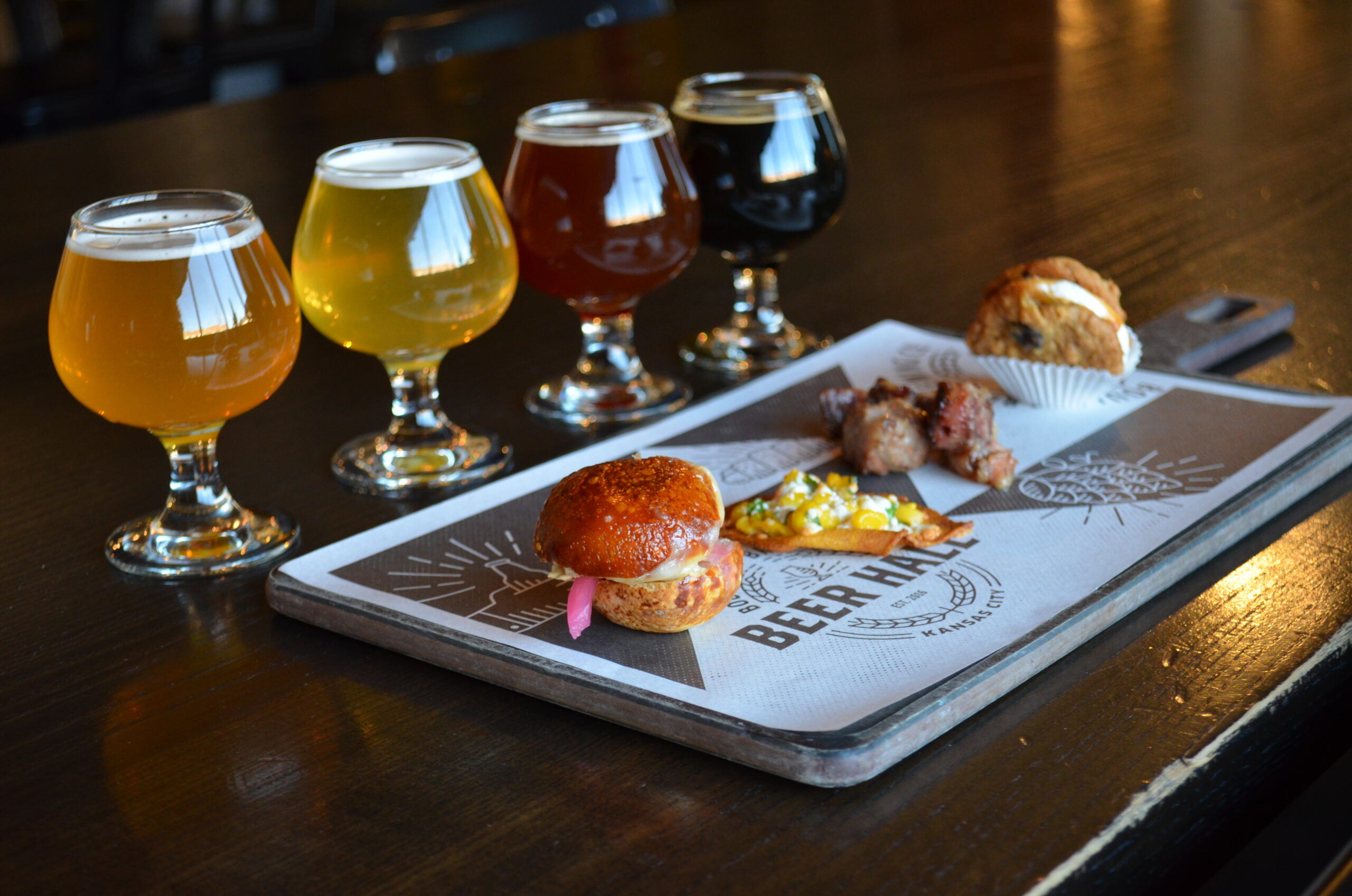 A flight of four beers in stemmed glasses served at Boulevard Brewing Co. Tours alongside a wooden board with a slider, a small taco, two pieces of meat, and a muffin.