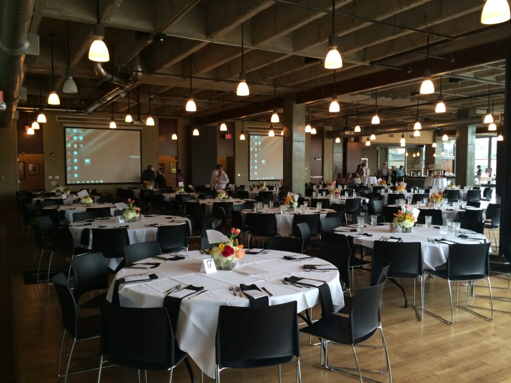 A spacious banquet hall with round tables covered in white tablecloths, each set with black napkins, utensils, and floral centerpieces. People are preparing the room, and projector screens are visible.