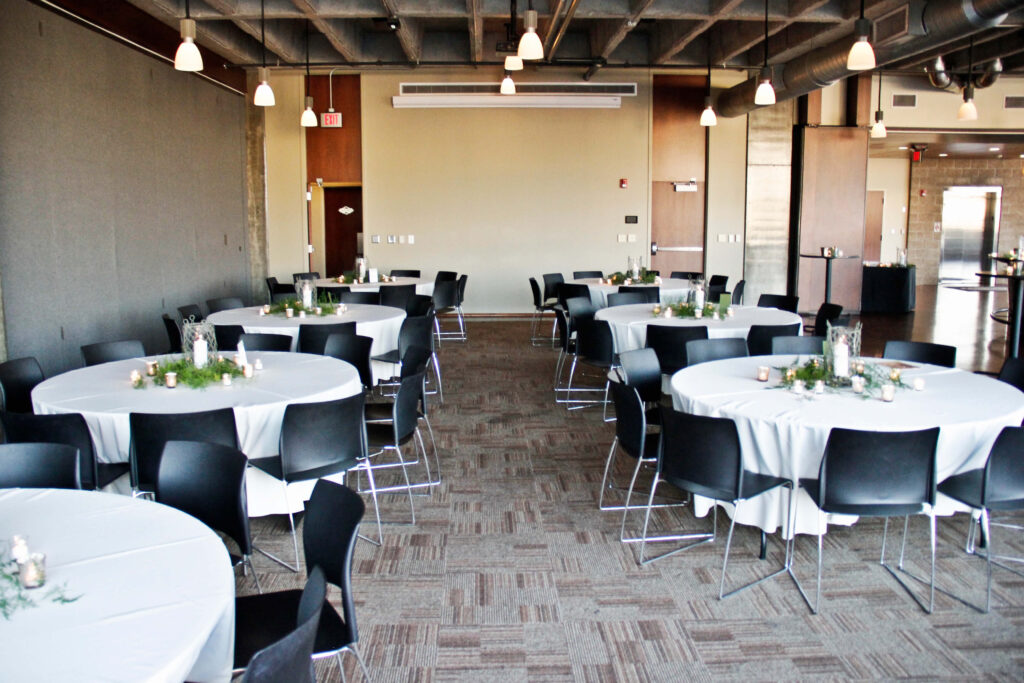 A banquet hall with round tables covered in white tablecloths, black chairs arranged around each table, and small green centerpieces. The room has carpeted floors, pendant lights, and a modern ceiling with exposed ducts.