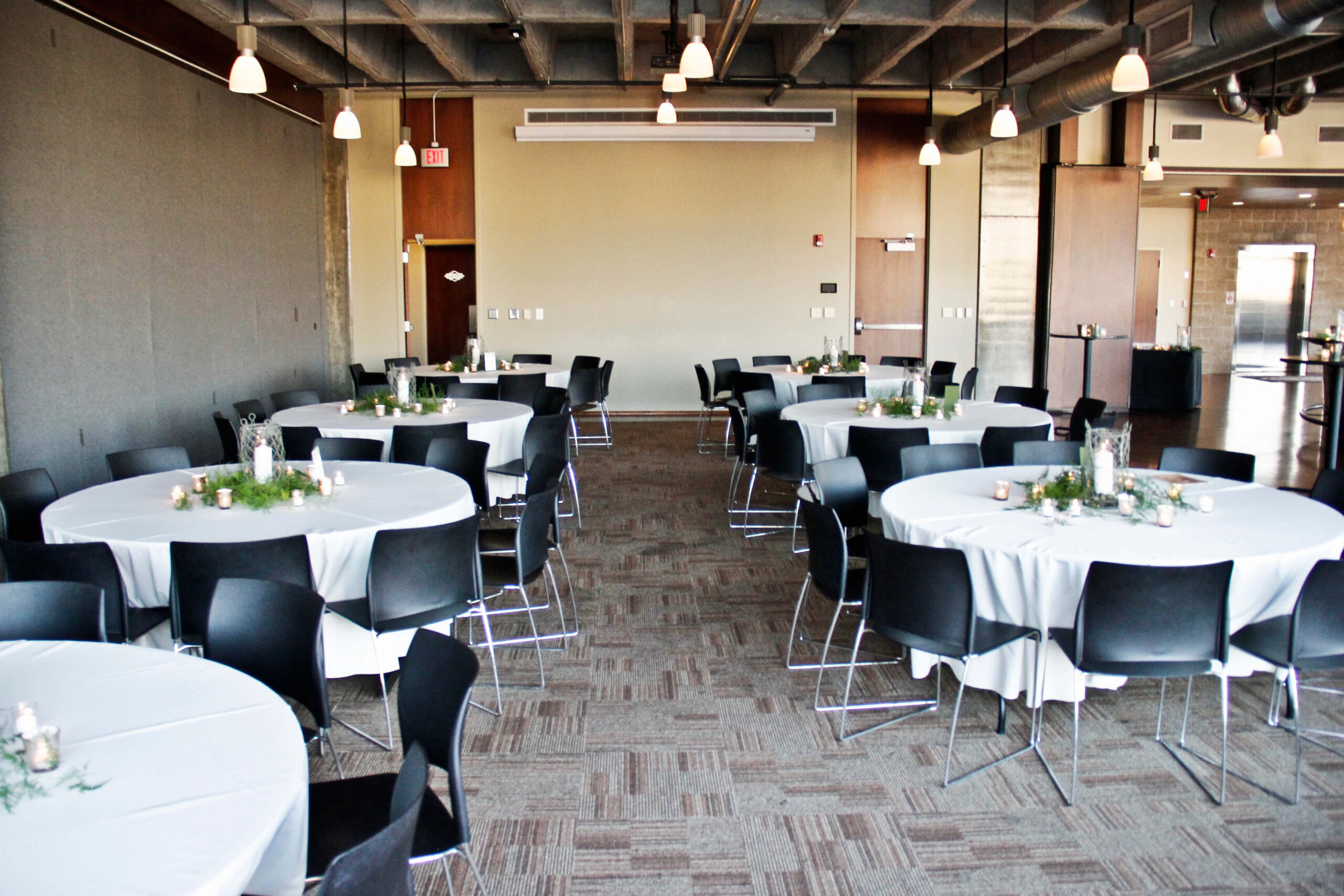 A banquet hall with round tables covered in white tablecloths, black chairs arranged around each table, and small green centerpieces. The room has carpeted floors, pendant lights, and a modern ceiling with exposed ducts.