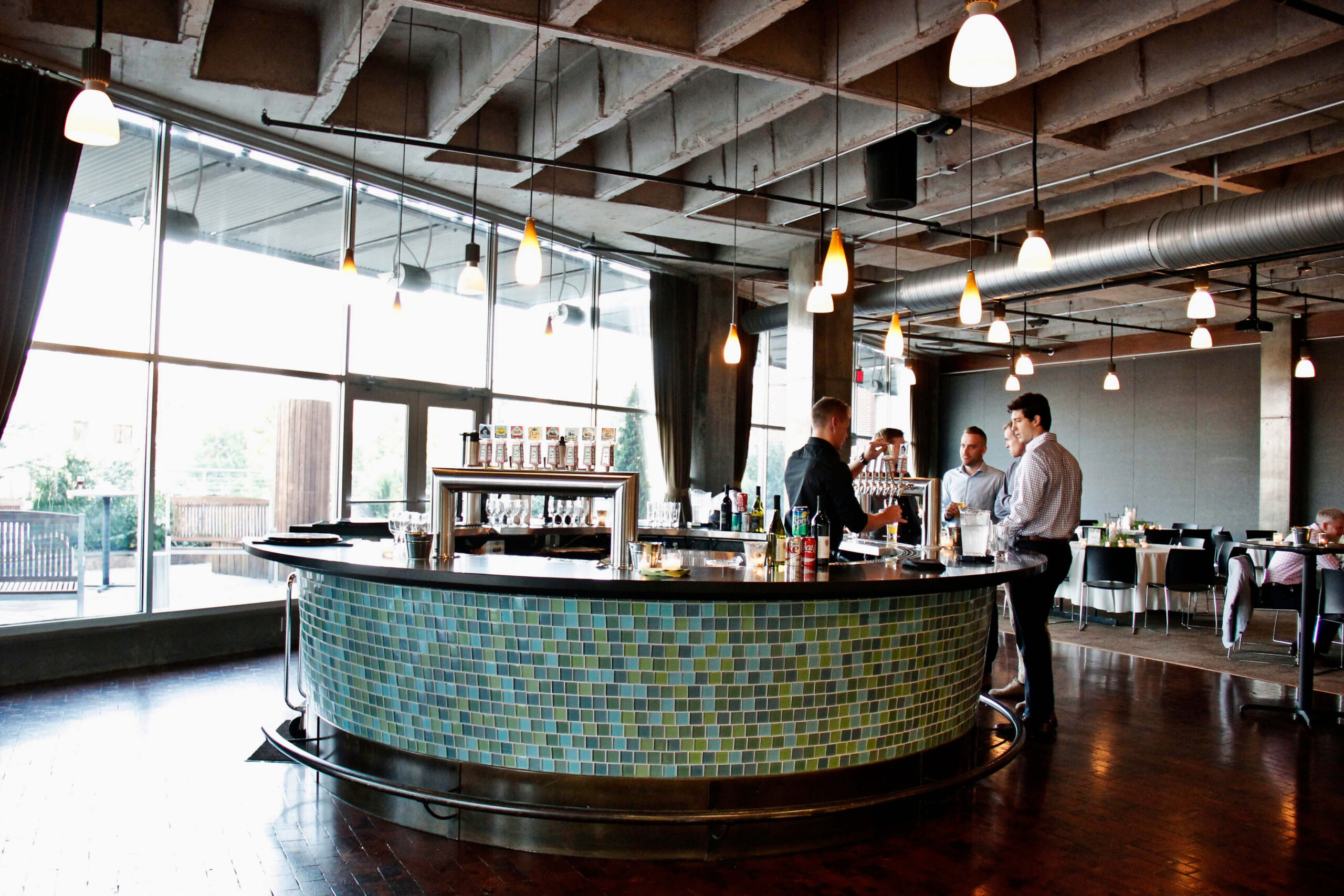 A modern bar with a circular counter covered in blue and green tiles, surrounded by people talking. Large windows let in natural light, and pendant lights hang from the exposed ceiling.