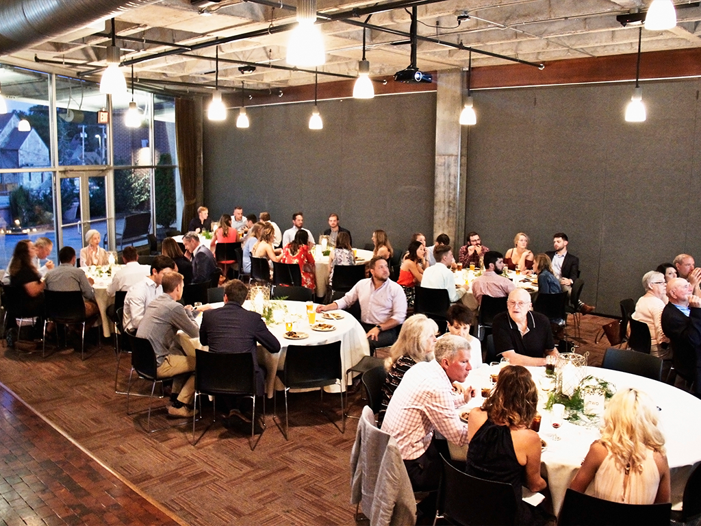 A large group of people seated around round tables at an indoor event. The ambiance is warm, with dim lighting. People are engaged in conversation, enjoying food and drinks. Decorations include small floral centerpieces on the tables.