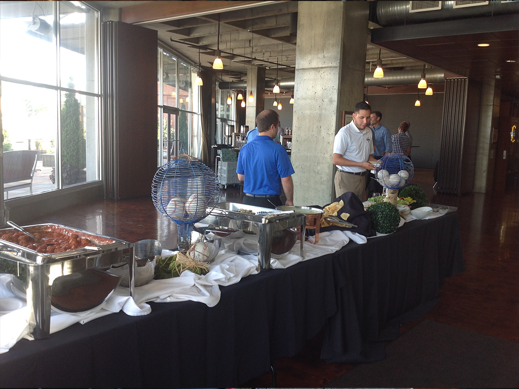 A buffet table is set up with various chafing dishes and decorative items. People in casual attire are serving themselves. The room has large windows, wooden floors, and industrial-style columns. Hanging lights illuminate the area.