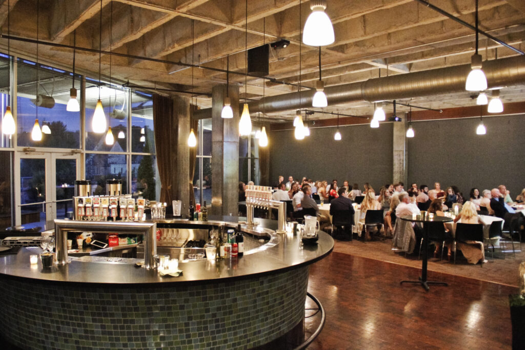 A modern, stylish restaurant with a large circular bar in the foreground, pendant lights hanging from the ceiling, and groups of people dining at round tables in the background.