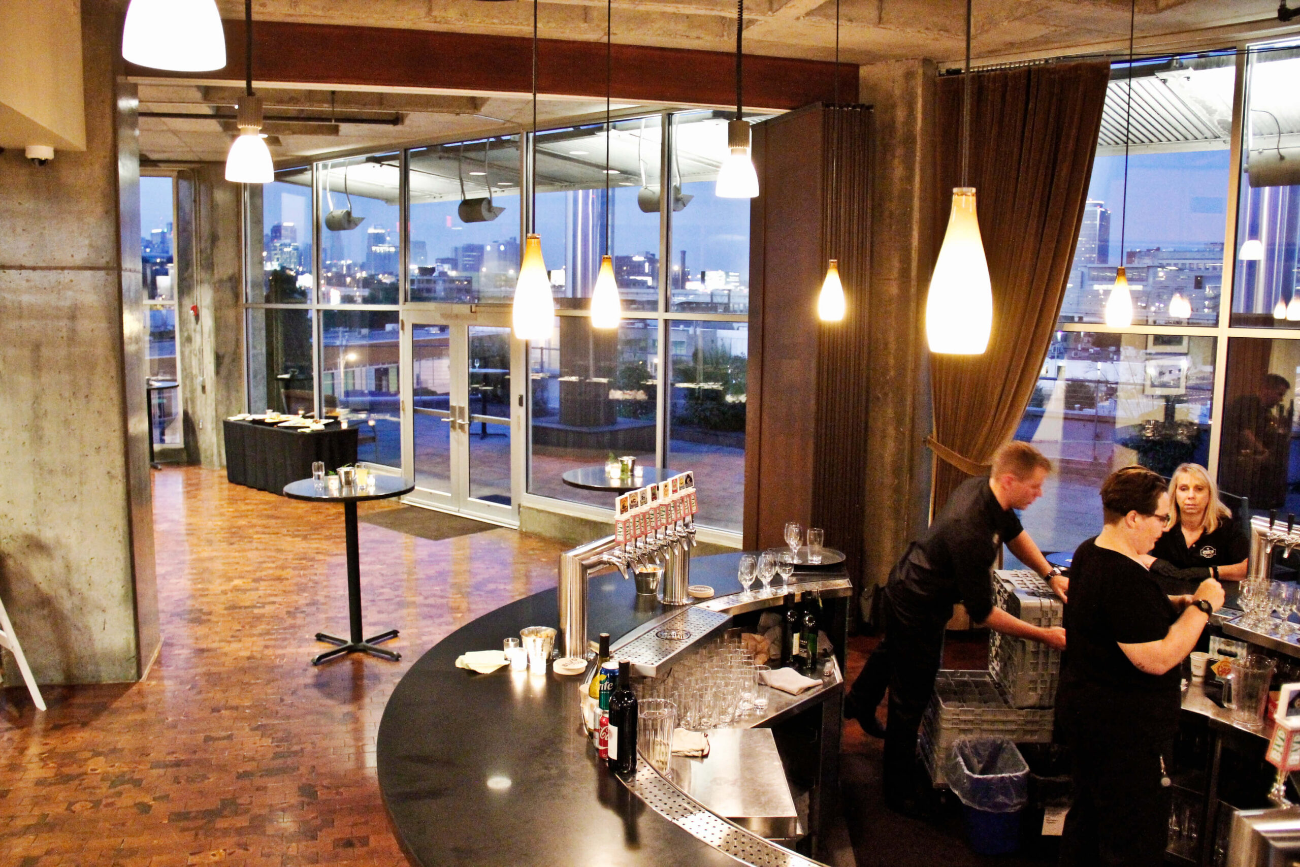 Modern bar with hanging lights, a curved counter, and large windows overlooking a cityscape at dusk. Three bartenders prepare drinks behind the bar. A high table with snacks is set up in the background.
