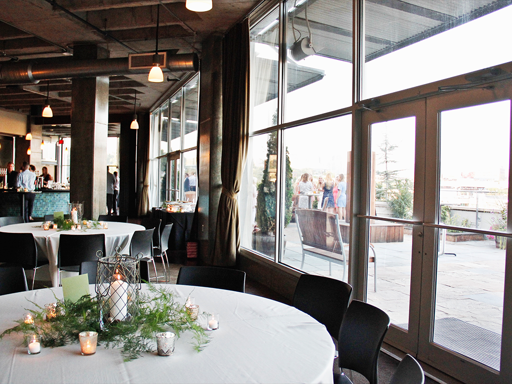 A bright, modern event space with round tables covered in white tablecloths and decorated with greenery and candles. Large windows and glass doors reveal a patio with people mingling in the background.