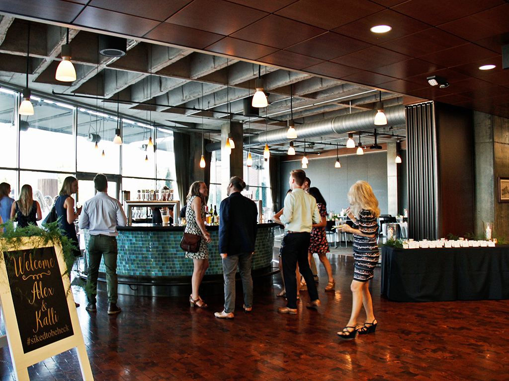 A group of people mingles at a modern indoor event space with a central bar, surrounded by hanging lights and wooden flooring. A sign welcomes guests to the event, and candles are arranged on a table to the right.