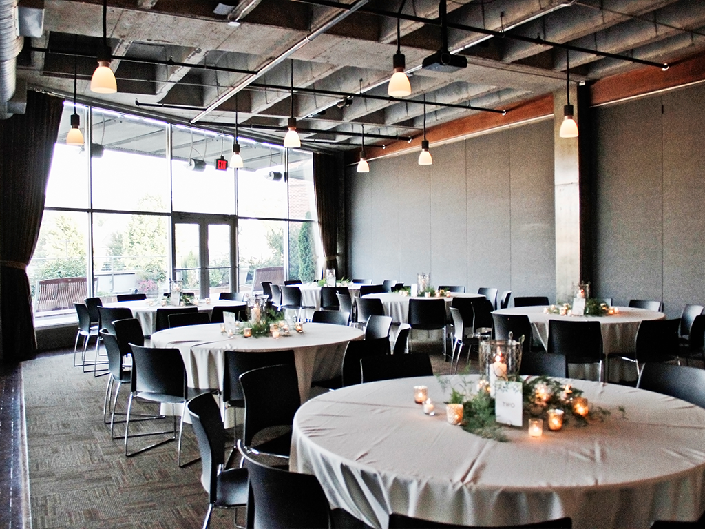 A spacious event room with round tables covered in beige tablecloths, decorated with candles and greenery. Black chairs are arranged around the tables. Large windows let in natural light, and modern pendant lights hang from the ceiling.