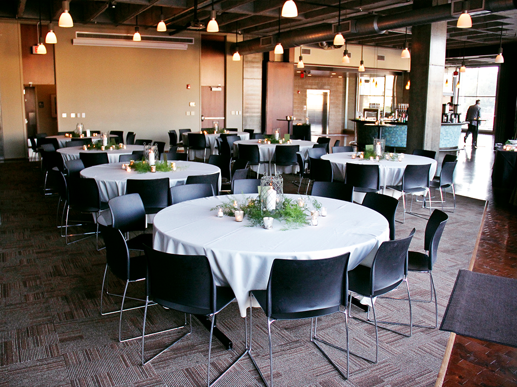 A decorated banquet hall with round tables covered in white tablecloths and surrounded by black chairs. Each table has a centerpiece with greenery and candles. Warm lighting hangs from the ceiling, creating a cozy ambiance.