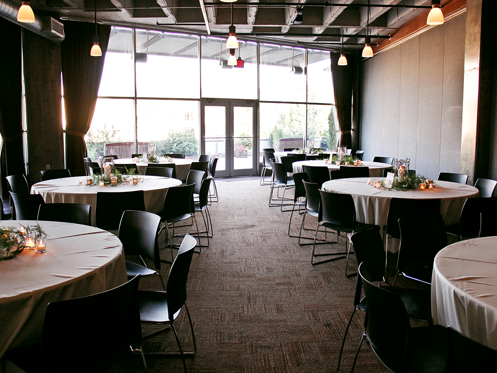 A banquet hall with round tables covered in white tablecloths, arranged with small floral centerpieces and surrounded by black chairs. Large windows and glass doors in the background allow natural light to fill the room.