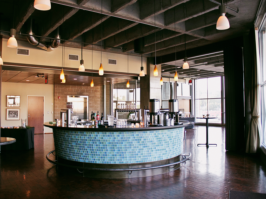 Industrial-style cafe with a circular counter covered in blue mosaic tiles. Pendant lights hang from the exposed ceiling, and large windows illuminate the space. Tables and chairs are scattered around the polished wooden floor.