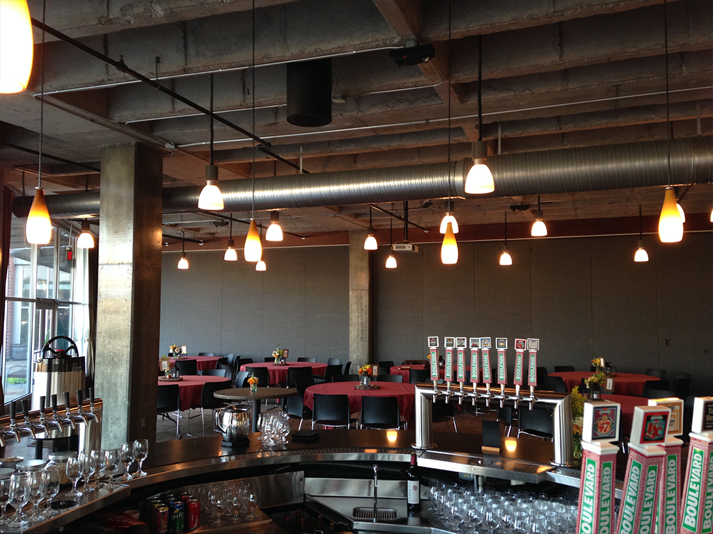 A modern, industrial-style bar with a concrete ceiling, exposed ducts, and hanging lights. The bar features taps, glasses, and red-topped tables with chairs in the background. Soft lighting creates a warm ambiance.