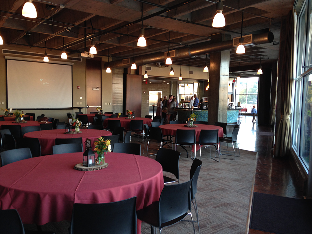 Spacious event hall with round tables covered in red tablecloths, black chairs, and small floral centerpieces. A large projector screen is at the front, and several ceiling lights illuminate the room. Appears calm and ready for guests.